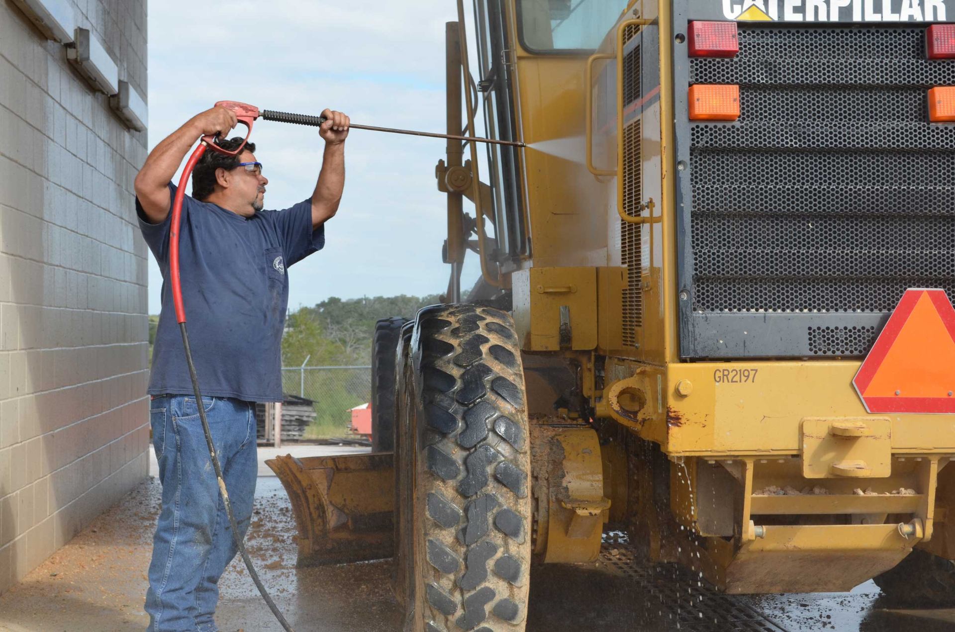 Worker pressure washing a Caterpillar motor grader to remove caked-on mud and grime using Enzo's construction equipment detergents